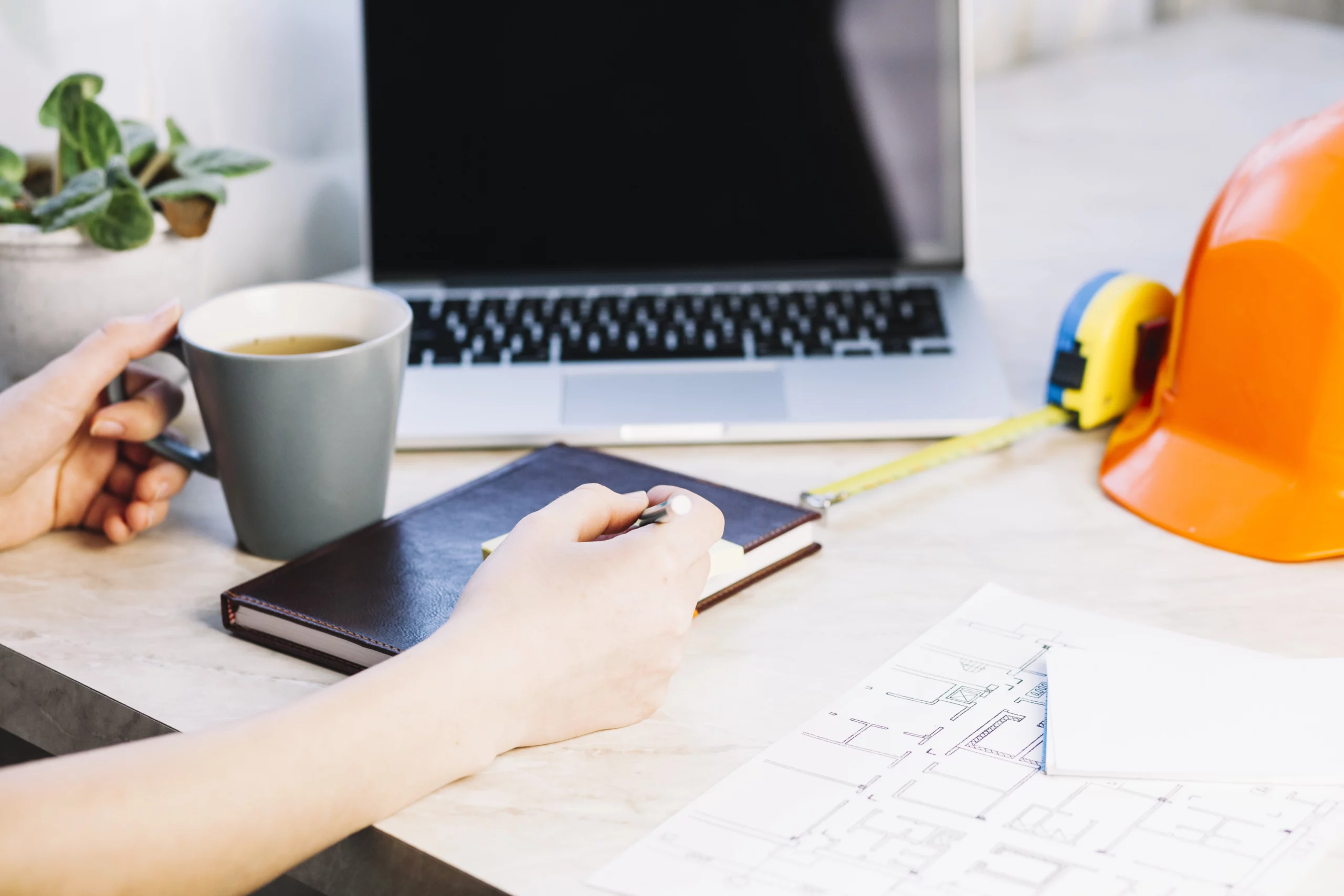 Artisan travaillant avec un ordinateur portable, un carnet de notes, une tasse de café et un plan de chantier sur un bureau