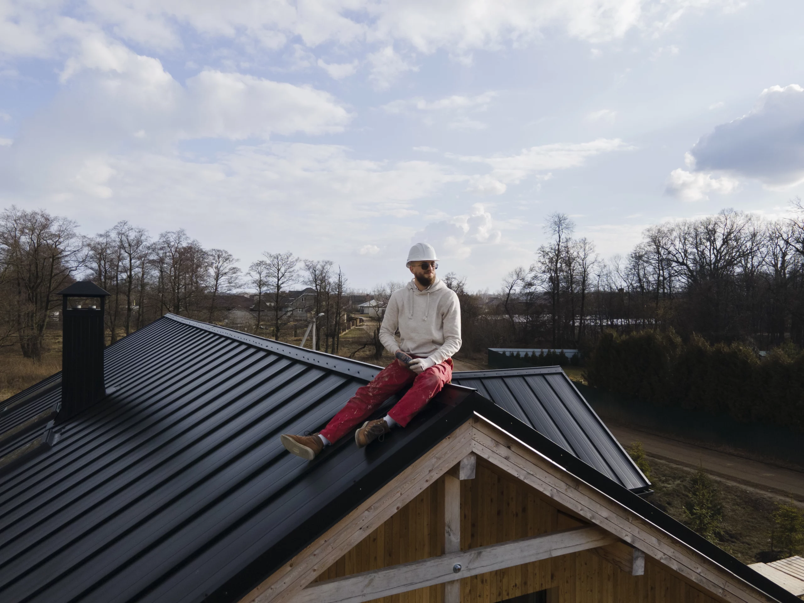 Couvreur assis sur un toit en métal noir d'une maison en bois, portant un casque de sécurité.