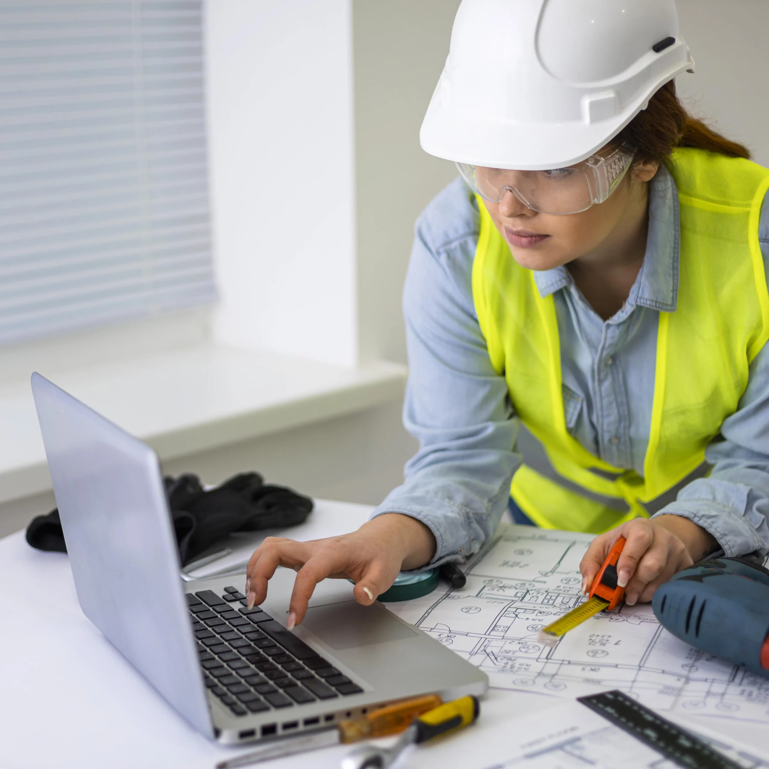 Artisan portant un casque de protection et un gilet de sécurité, travaillant sur un ordinateur portable pour créer un devis tout en consultant un plan de chantier.