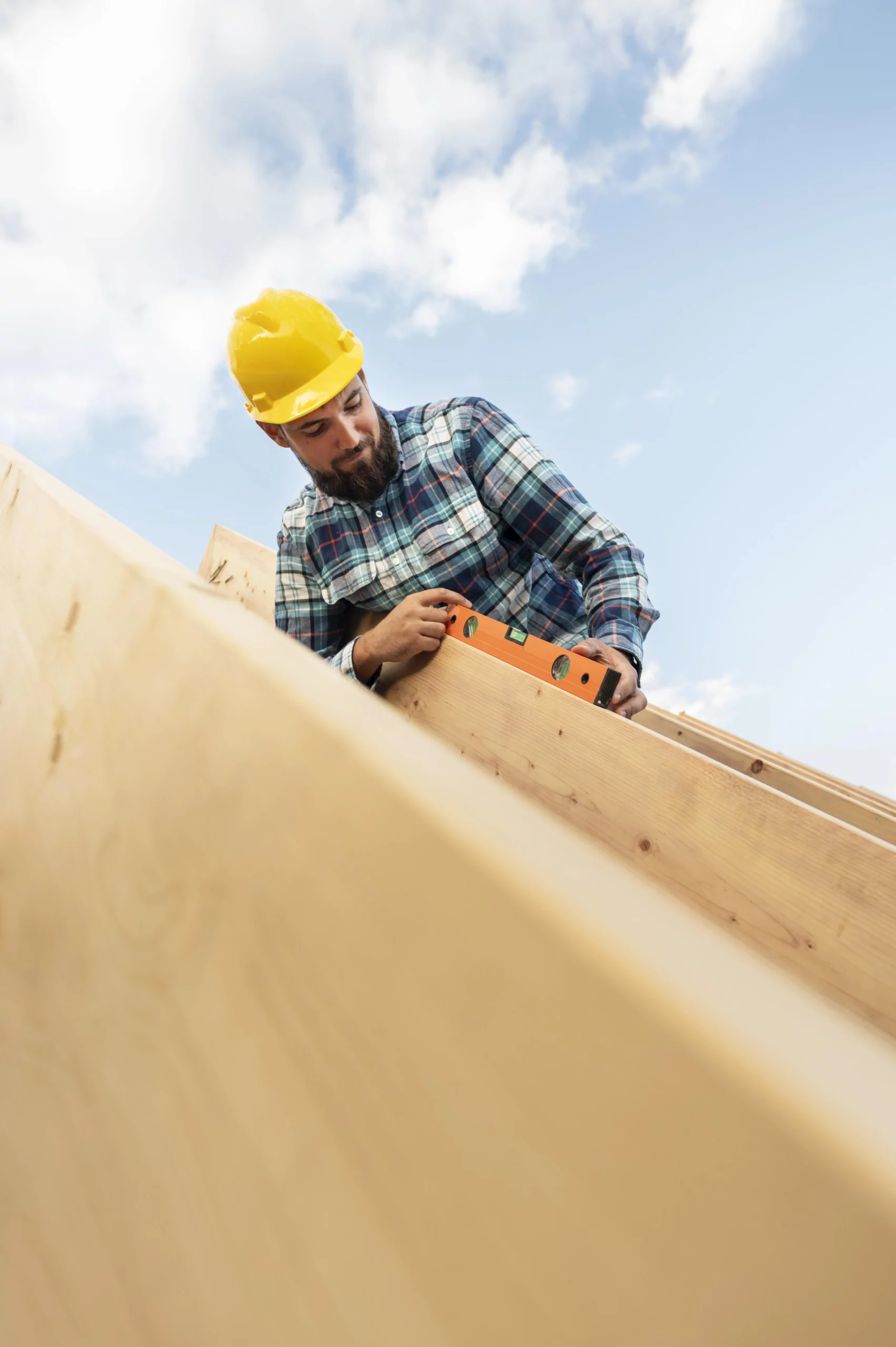 Charpentier portant des pièces de bois sur un chantier, vêtu d'une chemise à carreaux et d'un casque de protection.